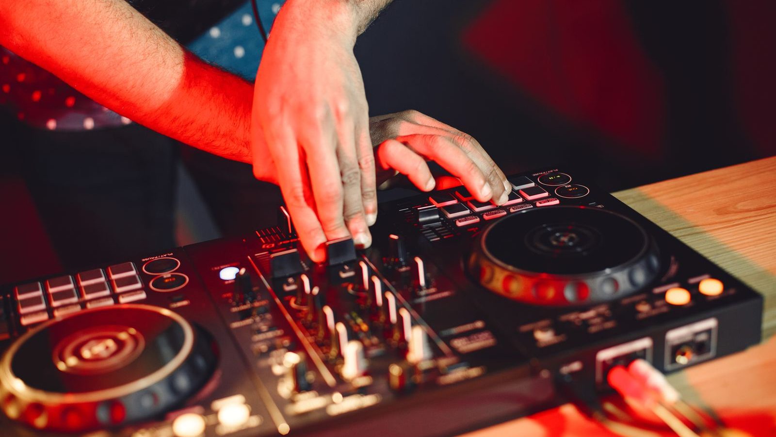 A DJ's hands working the jog wheels and pads of a controller under warm red light