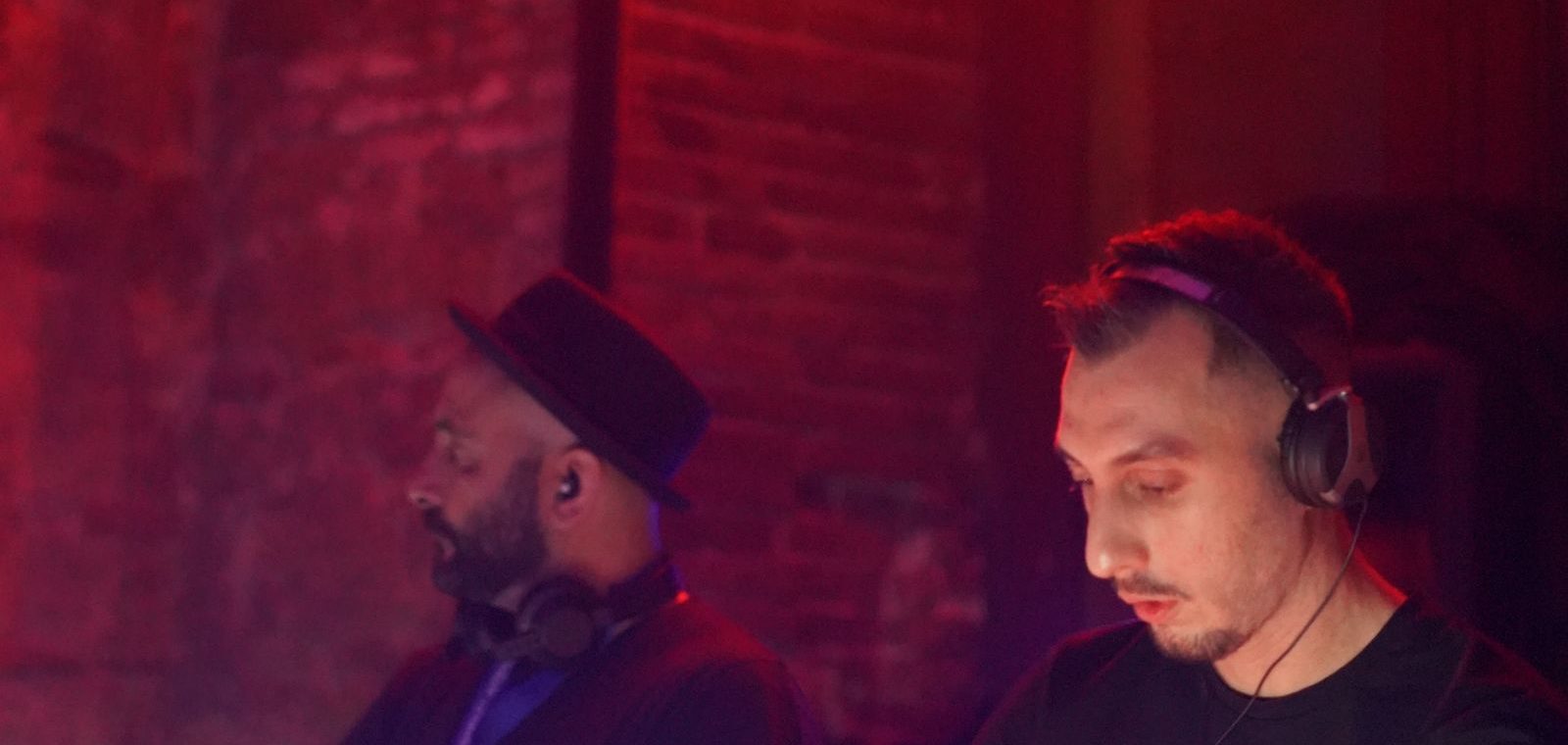 Two DJs playing a set on a full booth setup, lit under warm red club lighting