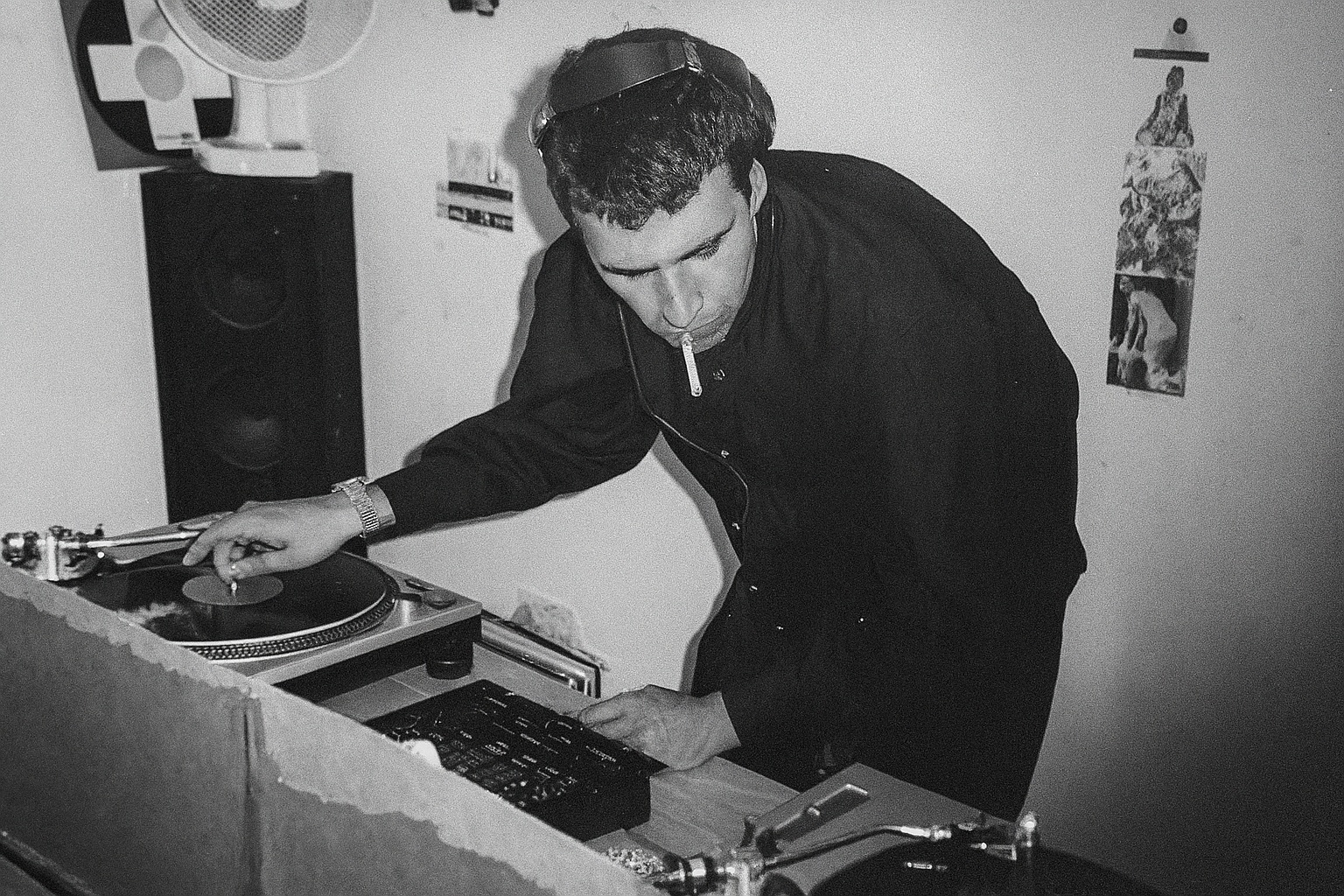 John behind a pair of Technics turntables in the 1990s, cueing a record with a cigarette in his mouth.